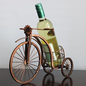 Bronze finish metal wine holder shaped like a bicycle, designed to display one wine bottle and two wine glasses in a charming and functional way, perfect for kitchen islands, dining rooms, wine stations, picnics, or outdoor entertaining, made from durable metal for long lasting use, serving as unique wine decor and a meaningful gift for weddings, anniversaries, housewarmings, holidays, and wine lovers who enjoy shared experiences