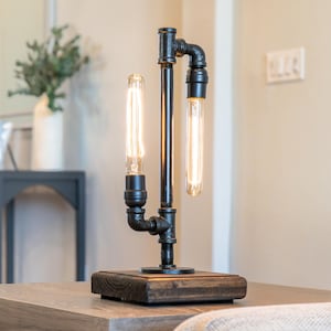 May include: A table lamp constructed from black metal plumbing pipes and fittings, featuring a rectangular dark wood base and two vertical Edison-style light bulbs that emit a warm glow, one positioned facing upward and one facing downward.