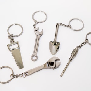 May include: Five silver keychains with different tool designs: a saw, a shovel, a wrench, a screwdriver, and a wrench. Each keychain has a silver ring.