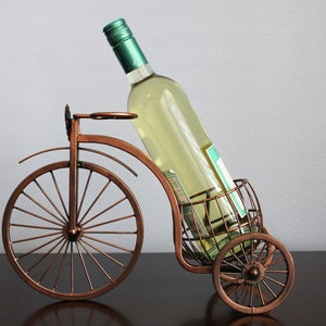 Decorative metal wine and glass holder finished in bronze and shaped like a bicycle, designed to securely hold one wine bottle and two glasses, ideal for kitchen displays, dining rooms, wine stations, and outdoor gatherings, crafted from durable metal for long term use, combining creativity and function, and making a unique wine decor accent or a meaningful gift for weddings, anniversaries, holidays, housewarmings, and wine loving couples