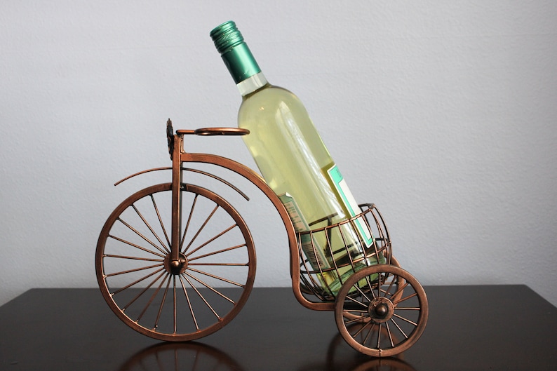 Decorative metal wine and glass holder finished in bronze and shaped like a bicycle, designed to securely hold one wine bottle and two glasses, ideal for kitchen displays, dining rooms, wine stations, and outdoor gatherings, crafted from durable metal for long term use, combining creativity and function, and making a unique wine decor accent or a meaningful gift for weddings, anniversaries, holidays, housewarmings, and wine loving couples