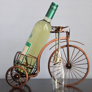 Unique metal wine and glass holder featuring a bronze finish and bicycle inspired design, created to hold one wine bottle and two glasses for easy sharing, ideal for kitchen counters, dining rooms, wine displays, and outdoor gatherings, crafted from sturdy metal for durability, and perfect as decorative wine holder decor, picnic wine display, or a thoughtful gift for weddings, anniversaries, holidays, and couples who appreciate functional yet creative home accents