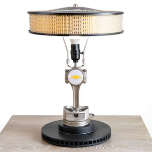May include: A table lamp made from a car engine piston, air filter, and a Chevrolet logo. The lamp has a black base and a black shade with a yellow air filter.