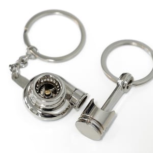 Puede incluir: Un llavero plateado con un turbocompresor y un encanto de biela. El turbocompresor tiene un acabado plateado y dorado y un diseño detallado. La biela es un diseño simple con un acabado plateado.
