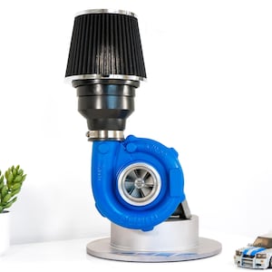 Op de afbeelding: Een blauwe turboladerlamp met een zwart luchtfilter en een zilveren voet. De lamp staat op een wit oppervlak met een kleine speelgoedauto op de voorgrond.