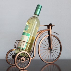 Decorative metal wine and glass holder finished in bronze and shaped like a bicycle, designed to securely hold one wine bottle and two glasses, ideal for kitchen displays, dining rooms, wine stations, and outdoor gatherings, crafted from durable metal for long term use, combining creativity and function, and making a unique wine decor accent or a meaningful gift for weddings, anniversaries, holidays, housewarmings, and wine loving couples