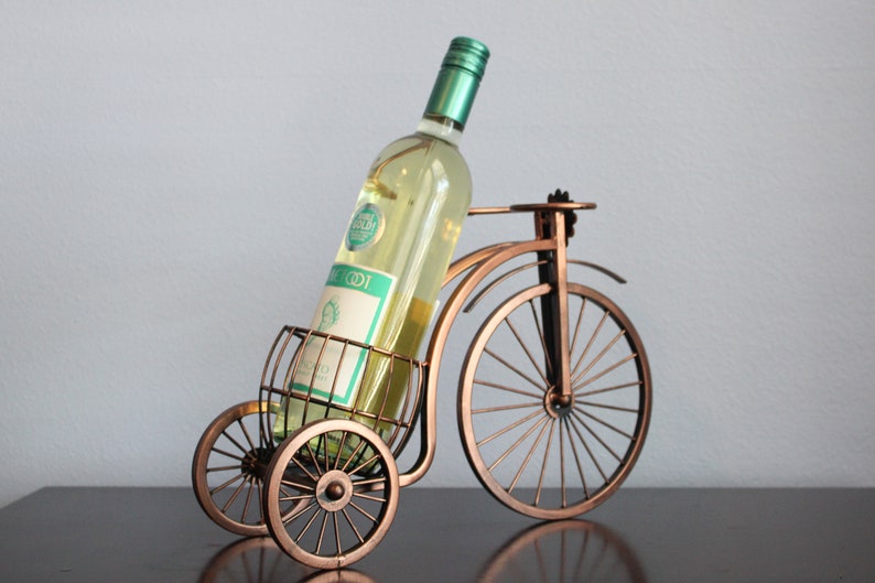 Decorative metal wine and glass holder finished in bronze and shaped like a bicycle, designed to securely hold one wine bottle and two glasses, ideal for kitchen displays, dining rooms, wine stations, and outdoor gatherings, crafted from durable metal for long term use, combining creativity and function, and making a unique wine decor accent or a meaningful gift for weddings, anniversaries, holidays, housewarmings, and wine loving couples