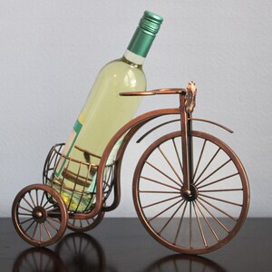 Unique bronze finish metal wine and glass holder shaped like a bicycle, designed to display one wine bottle and two glasses with charm and balance, ideal for kitchens, dining rooms, wine displays, and outdoor gatherings, crafted from durable metal for long lasting use, offering both function and style, and making a thoughtful gift for couples, weddings, anniversaries, holidays, housewarmings, and anyone who enjoys creative wine decor