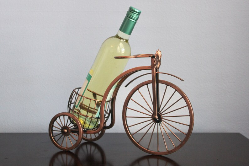 Unique bronze finish metal wine and glass holder shaped like a bicycle, designed to display one wine bottle and two glasses with charm and balance, ideal for kitchens, dining rooms, wine displays, and outdoor gatherings, crafted from durable metal for long lasting use, offering both function and style, and making a thoughtful gift for couples, weddings, anniversaries, holidays, housewarmings, and anyone who enjoys creative wine decor