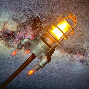 May include: A unique, industrial-style lamp shaped like a rocket, with a silver metal body and a caged light fixture. The lamp has three small, orange-red flame-like accents and a long, dark brown pole. The background is a starry night sky.