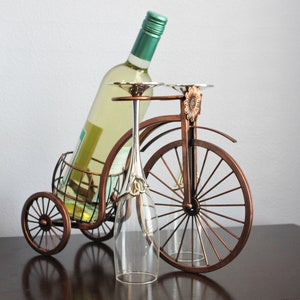 Metal wine bottle holder with a bronze finish and bicycle inspired design, crafted to hold one wine bottle and two wine glasses for shared enjoyment, perfect for kitchen counters, dining rooms, wine stations, picnics, and outdoor entertaining, built from durable metal for lasting quality, serving as unique wine decor and a meaningful gift for weddings, anniversaries, holidays, housewarmings, and wine lovers who value thoughtful design