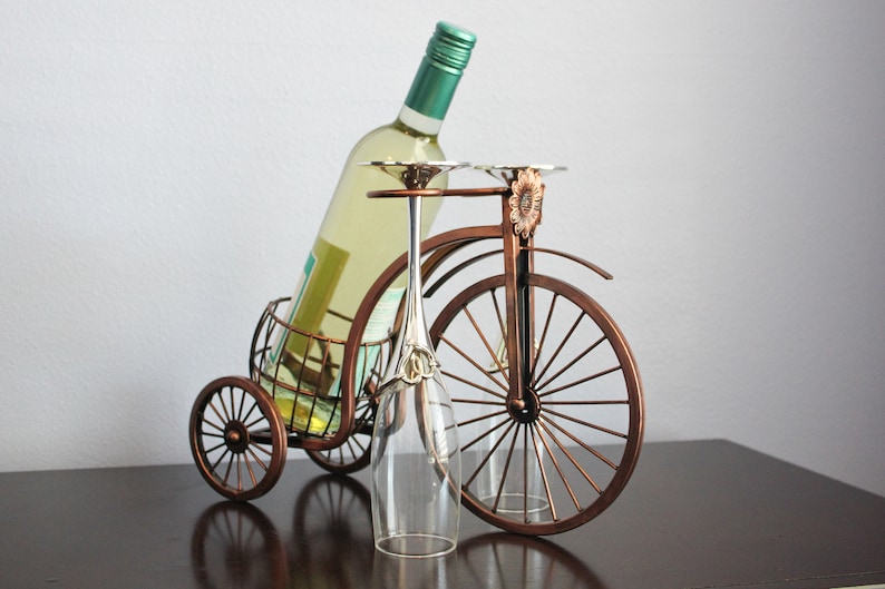 Metal wine bottle holder with a bronze finish and bicycle inspired design, crafted to hold one wine bottle and two wine glasses for shared enjoyment, perfect for kitchen counters, dining rooms, wine stations, picnics, and outdoor entertaining, built from durable metal for lasting quality, serving as unique wine decor and a meaningful gift for weddings, anniversaries, holidays, housewarmings, and wine lovers who value thoughtful design