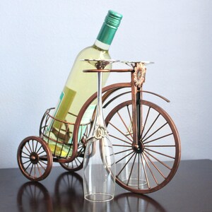 Metal wine and glass holder finished in a rich bronze tone, designed to hold one wine bottle and two wine glasses in a creative bicycle shaped display, perfect for kitchen counters, dining rooms, wine stations, and outdoor gatherings, offering a balance of charm and function, crafted from durable metal for long lasting use, ideal as unique wine decor, picnic wine holder, or a thoughtful gift for couples, weddings, anniversaries, and housewarming celebrations