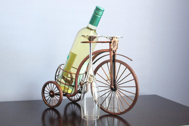 Metal wine and glass holder finished in a rich bronze tone, designed to hold one wine bottle and two wine glasses in a creative bicycle shaped display, perfect for kitchen counters, dining rooms, wine stations, and outdoor gatherings, offering a balance of charm and function, crafted from durable metal for long lasting use, ideal as unique wine decor, picnic wine holder, or a thoughtful gift for couples, weddings, anniversaries, and housewarming celebrations