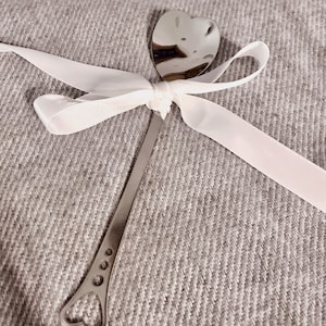 Op de afbeelding: Een roestvrijstalen lepel met een hartvormige kom en handvat. De handgreep heeft drie kleine cirkelvormige uitsparingen en een hartvormig uiteinde. Een wit satijnen lint is om de lepel bij de kom gebonden.