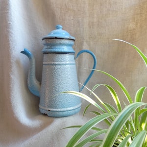 Può includere: Una caffettiera in smalto blu chiaro con una finitura testurizzata e un bordo nero. La caffettiera ha un beccuccio e un manico. La caffettiera è appoggiata su uno sfondo di lino bianco.