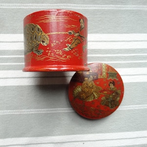 Op de afbeelding: Rode en gouden cilindrische doos met een deksel. De doos is versierd met een geschilderd tafereel van een schildpad en twee figuren. Het deksel is versierd met een geschilderd tafereel van twee figuren in traditionele Japanse kleding.