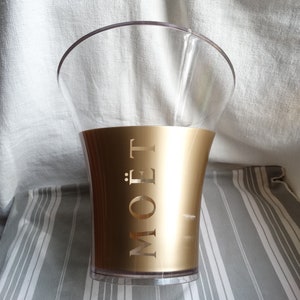 May include: A gold champagne bucket with the word "MOET" embossed on the side. The bucket is made of clear plastic and has a wide, flared opening.