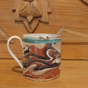 Puede incluir: Una taza de cerámica vintage con un asa blanca y un diseño veteado en tonos marrón, negro y crema. El borde presenta dos rayas verdes. La taza está sobre un fondo de madera.