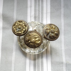 May include: Three antique brass knobs with intricate floral patterns, displayed on a clear glass object against a striped backdrop. The knobs exhibit a detailed, aged look, indicative of vintage craftsmanship. The floral designs are the main feature.