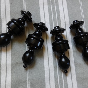 Puede incluir: Cuatro remates de madera negros con base bulbosa y parte superior redondeada. Los remates están dispuestos sobre una tela a rayas grises y blancas. Cada remate tiene un pequeño pomo redondo en la parte inferior.