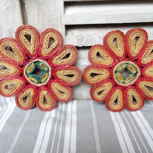 Può includere: Due sottopentola a forma di fiore intrecciati in rosso, giallo e verde. I sottopentola sono fatti di paglia e hanno una forma circolare con un foro al centro.