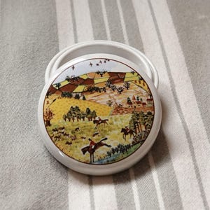 May include: A round ceramic trinket box with a white lid and a colourful scene of a hunt in a countryside setting. The scene features riders on horseback, hounds, and a small village in the distance.