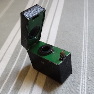 Peut inclure: Un petit encrier en cuir noir avec un intérieur vert et une plume en métal. Le couvercle est ouvert, révélant l'encrier.