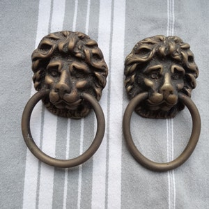 Puede incluir: Dos pomos de puerta de latón antiguo con forma de cabeza de león con tiradores de anillo. Las cabezas de león están detalladas con pelo texturizado y tienen una expresión realista.