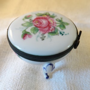 May include: A small, white porcelain trinket box with a floral design. The lid is decorated with a pink rose and green leaves. The box has three legs and a black metal hinge.