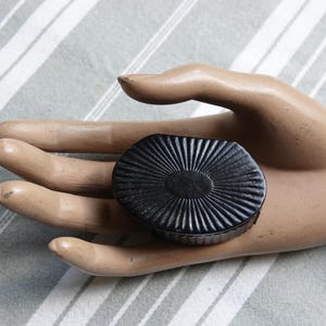 以下が含まれることがあります： 黒い楕円形のコンパクトで、蓋にサンバーストのデザインがあります。 ストライプの背景に対して、マネキンの手でコンパクトが保持されています。 コンパクトの表面は、テクスチャ加工されたリブ模様です。