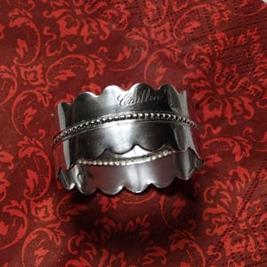 May include: A silver napkin ring with a scalloped edge and a beaded band. The name "Caitlin" is engraved on the top edge. The napkin ring is on a red patterned surface.