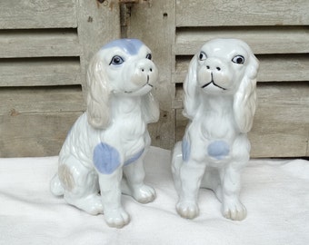 May include: Two porcelain figurines of dogs, both are white with blue spots. The dogs are sitting with their heads tilted up. The figurines are on a white surface.