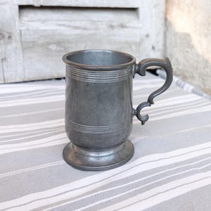 Puede incluir: Un tanque de peltre con un cuerpo redondeado y un asa. El tanque tiene un acabado liso, gris y está decorado con una serie de líneas horizontales alrededor del cuerpo.