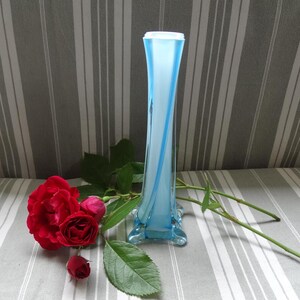 May include: A blue and white glass vase with a twisted design. The vase is sitting on a striped tablecloth with a red rose and green leaves.