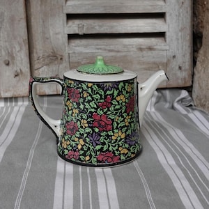 May include: A white teapot with a black and green floral pattern. The teapot has a green lid and a black rim.