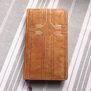 May include: A brown leather-bound book with gold detailing. The cover has a cross design with leaf patterns. A purple ribbon bookmark is visible. The book is on a grey and white striped surface.