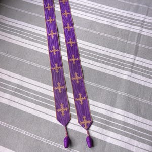 May include: Two purple liturgical stoles with gold crosses and trim. Each stole has a matching purple tassel at the bottom. The stoles are lying on a gray and white striped surface.