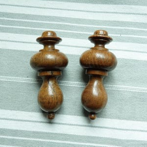 Puede incluir: Dos remates de madera con forma bulbosa y parte superior redondeada. Los remates son de color marrón oscuro y tienen un acabado liso y pulido.