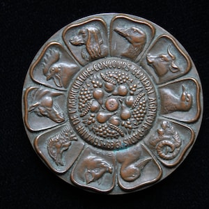 Puede incluir: Medalla de color bronce con diseño circular. La medalla presenta ilustraciones de animales alrededor del perímetro, incluyendo un perro, un pato, una vaca y un carnero. El centro tiene un diseño de frutas y verduras, con texto en francés: "CONCOURS GENERAL AGRICOLE PARIS 1961".