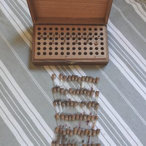 May include: A rectangular wooden storage box with a hinged lid and a grid of circular holes containing small metal pins, accompanied by a collection of numerous small, clear glass vials with cork stoppers, each holding various tiny contents.
