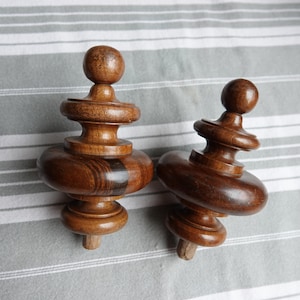May include: Two turned wooden finials in a dark brown hue. Each has a ball top, a series of rounded and curved sections, and a small peg at the base. They are displayed on a grey and white striped surface. The finials are likely for furniture.