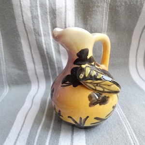 Puede incluir: Una pequeña jarra de cerámica amarilla con un diseño de abeja negro y amarillo. La jarra tiene un asa y un cuerpo redondeado.