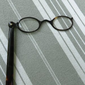 May include: Antique eyeglasses with oval lenses and a single handle. The frames are dark brown with a tortoiseshell pattern on the temples. The glasses are set against a gray and white striped background.