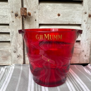 Könnte beinhalten: Ein transparenter roter Glaseiskübel mit dem goldenen Schriftzug G.H. MUMM auf der Vorderseite. Unter dem Text befindet sich ein kleines goldenes rechteckiges Emblem mit einem dünnen roten Rand. Der Eiskübel hat einen breiten Rand und integrierte Griffe.