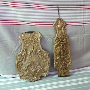 Könnte beinhalten: Zwei verzierte, goldfarbene Wandleuchter. Einer hat die Form eines Schildes mit floralen und Weizenähren-Details. Der andere ist ein hohes, schmales Design mit Trauben- und Sonnenblumenmotiven. Beide haben eine Vintage-Ästhetik.