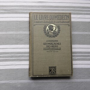 May include: A vintage French book, "LE LIVRE DU MEDECIN", with gold lettering and an emblem. The cover is a muted green, with text including "LES MALADIES DES-REINS". The book rests on a striped surface. Measurements are not available.