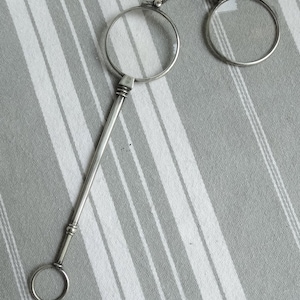 May include: Silver-toned magnifying glasses with a long handle and a circular ring at the end. The glasses are on a striped fabric background.