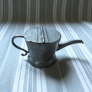 May include: A small, antique pewter oil can with a hinged lid and a curved spout. The handle is a curved shape. The can is a muted gray color and sits on a striped surface.