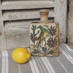 May include: Rectangular ceramic bottle with a bird and floral design. The bird is green with a long, brown tail. The bottle has a beige background with colorful flowers and leaves. A yellow lemon sits next to the bottle.